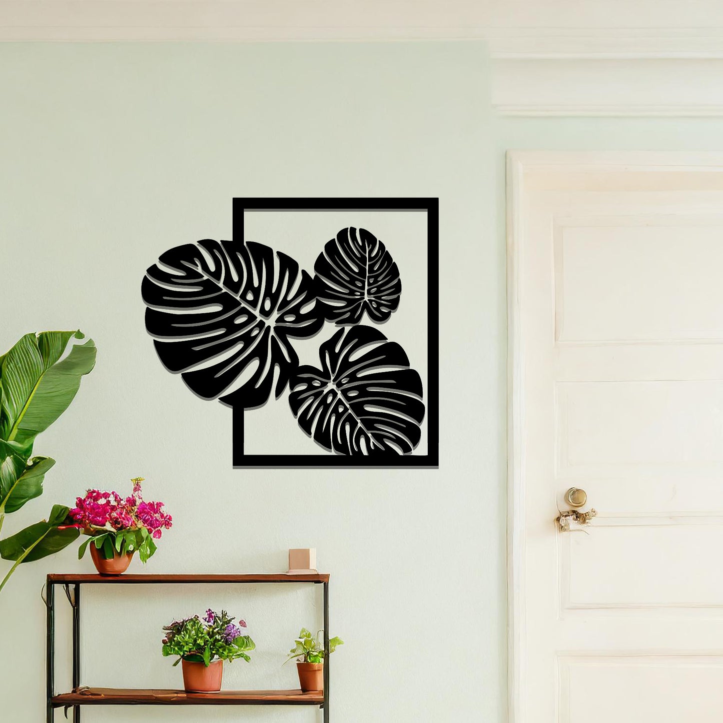 Metal wall art featuring a design of three large, detailed Monstera leaves arranged within a rectangular frame. The intricate cut-out pattern highlights the natural lines and veins of the leaves. The artwork is displayed against a neutral wall in a modern setting.
