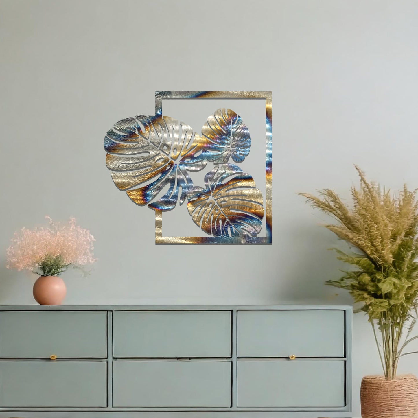 Metal wall art featuring a design of three large, detailed Monstera leaves arranged within a rectangular frame. The intricate cut-out pattern highlights the natural lines and veins of the leaves. The artwork is displayed against a neutral wall in a modern setting.