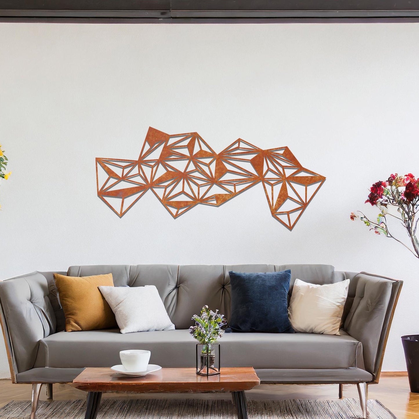 Metal wall art featuring an abstract geometric design with multiple interconnected triangular shapes, creating a dynamic, faceted pattern. The art is placed on a neutral wall within a modern room, complementing the vibrant decor with plants and furniture.