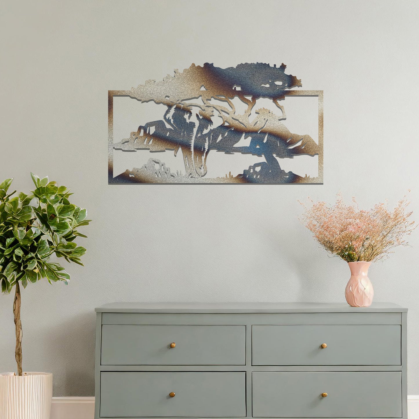 Metal wall art featuring an elephant silhouette under a tree with mountains in the background. The design is framed and placed on a neutral wall with contemporary furniture.