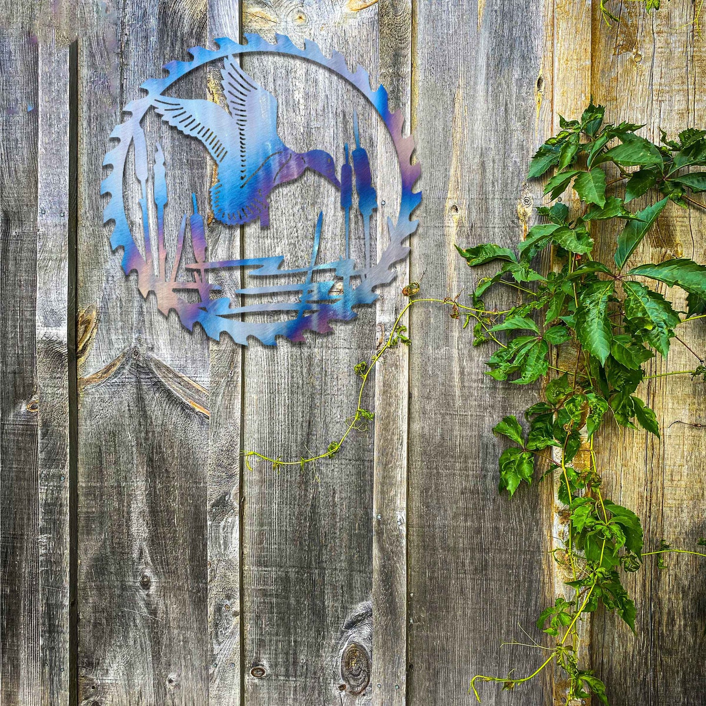 Metal garden decoration featuring a duck in flight over a pond surrounded by cattails, enclosed within a circular saw blade design, combining rustic charm and outdoor-inspired artistry to enhance garden or wall decor.