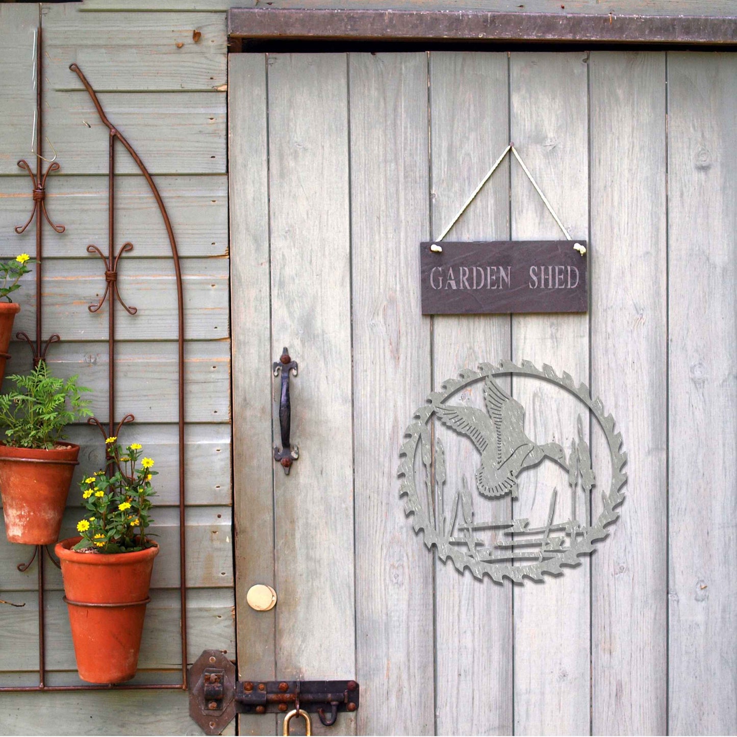 Metal garden decoration featuring a duck in flight over a pond surrounded by cattails, enclosed within a circular saw blade design, combining rustic charm and outdoor-inspired artistry to enhance garden or wall decor.