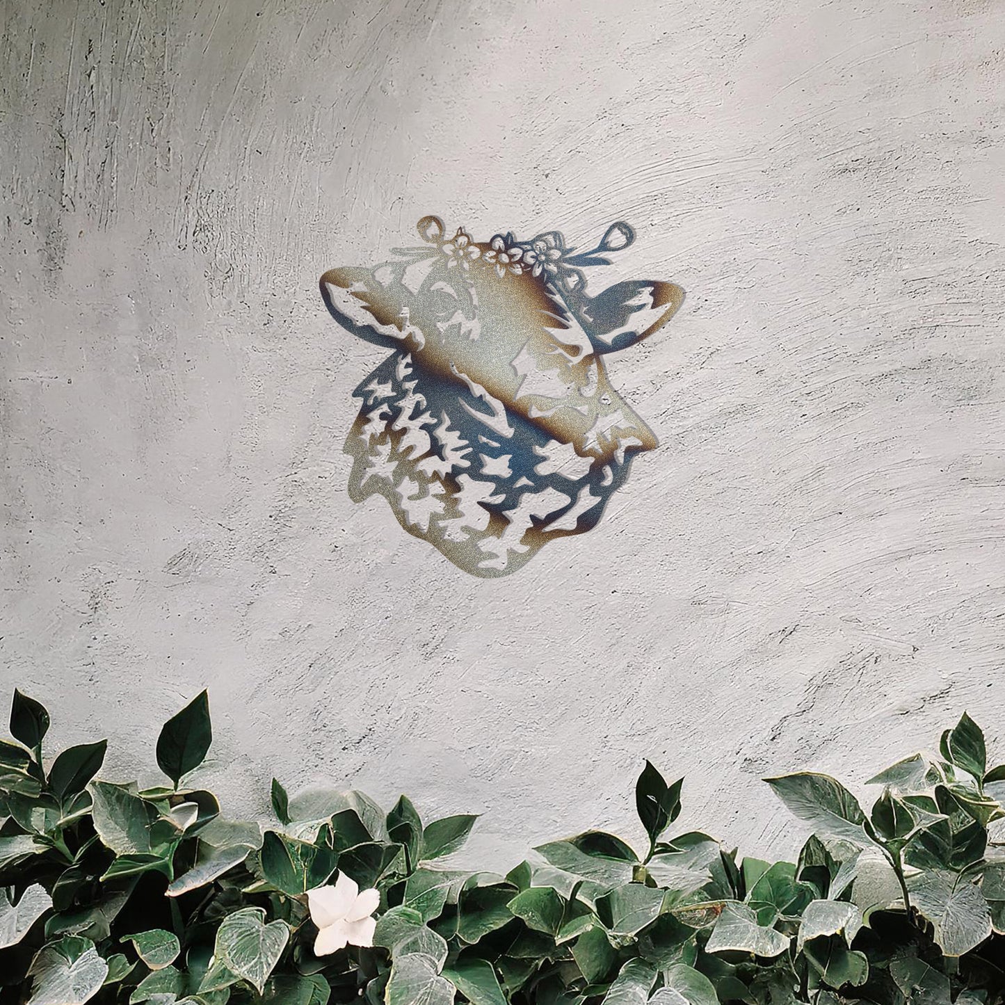 A metal wall art piece depicting a sheep adorned with a floral crown, exuding rustic charm and elegance. This intricate design brings a touch of countryside serenity to any modern or natural setting, complemented by its leafy surroundings.