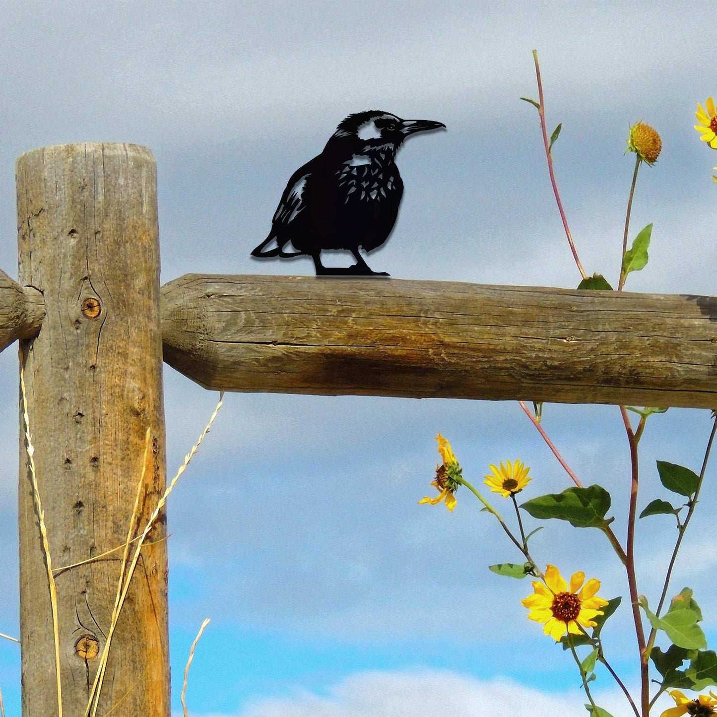 Metal wall art of a bird perched on a wooden fence post, featuring detailed wing and feather patterns. The bird is in a relaxed pose, surrounded by sunflowers, showcasing nature's beauty.