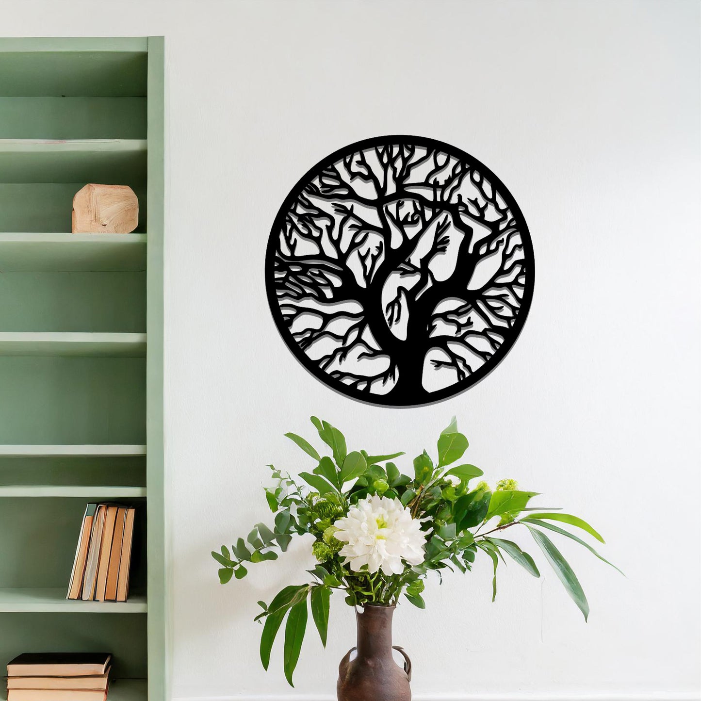 Metal wall art depicting a beautifully intricate tree design with twisting branches inside a circular frame. The artwork is placed on a neutral wall within a modern space, enhanced by the surrounding greenery and bookshelves.
