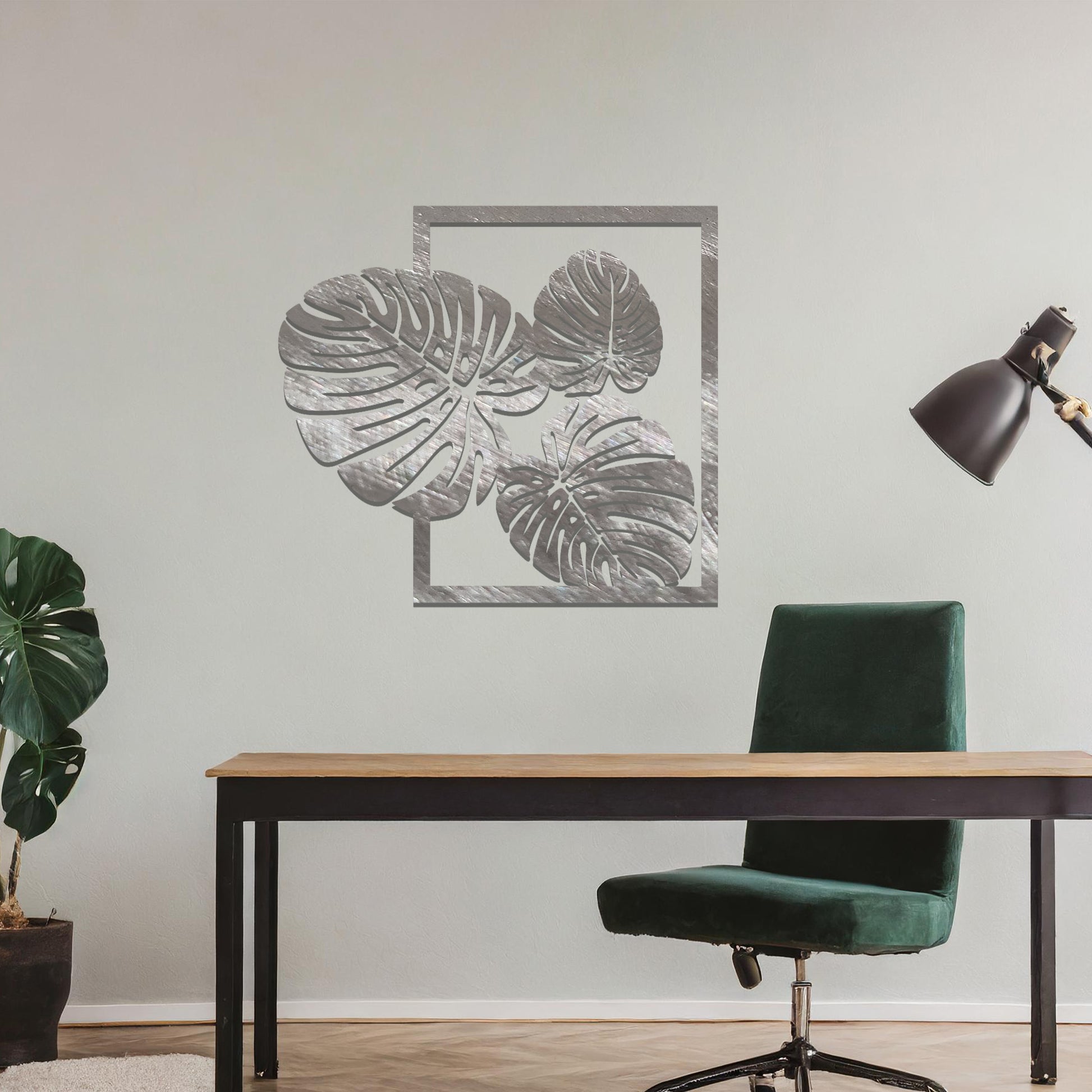 Metal wall art featuring a design of three large, detailed Monstera leaves arranged within a rectangular frame. The intricate cut-out pattern highlights the natural lines and veins of the leaves. The artwork is displayed against a neutral wall in a modern setting.