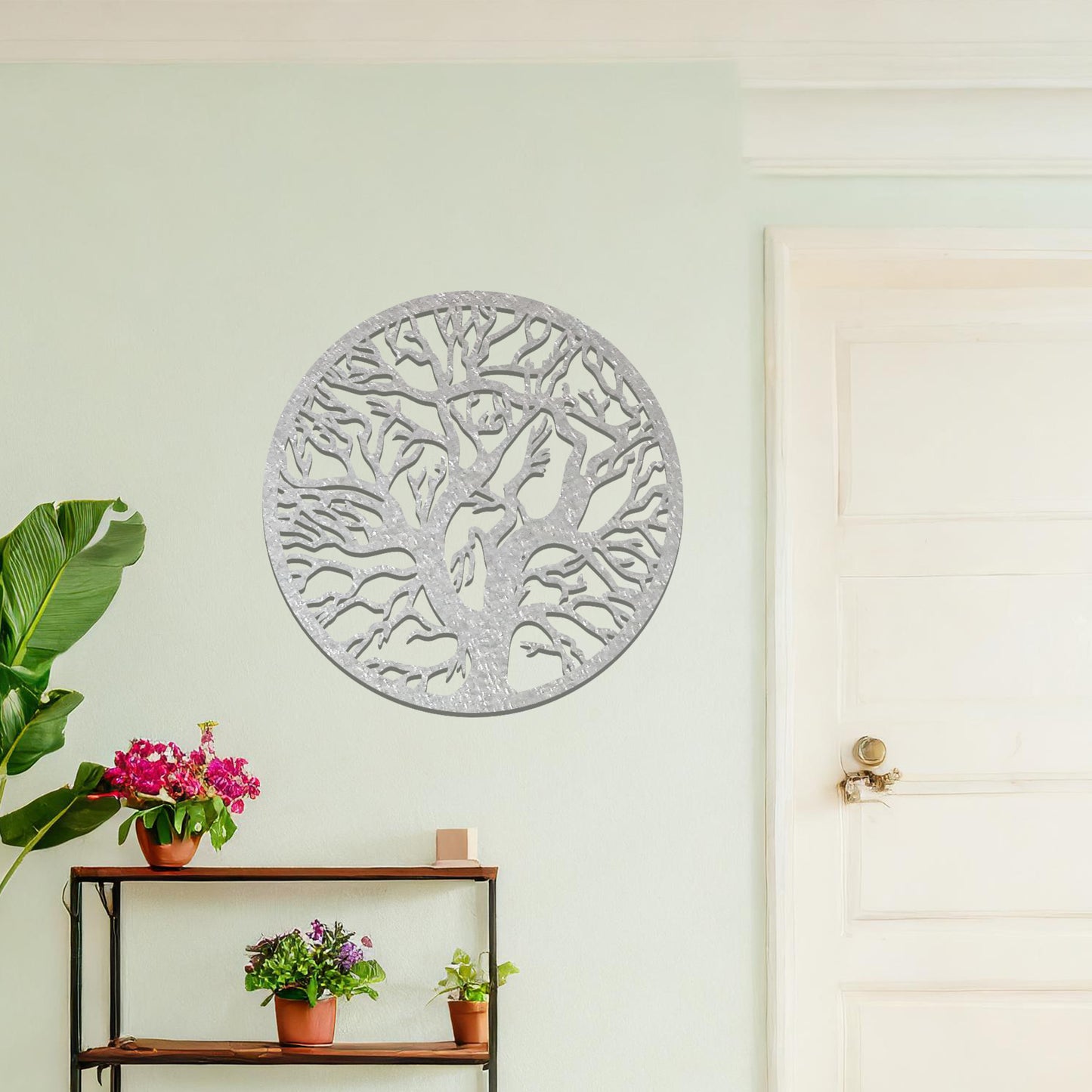 Metal wall art depicting a beautifully intricate tree design with twisting branches inside a circular frame. The artwork is placed on a neutral wall within a modern space, enhanced by the surrounding greenery and bookshelves.