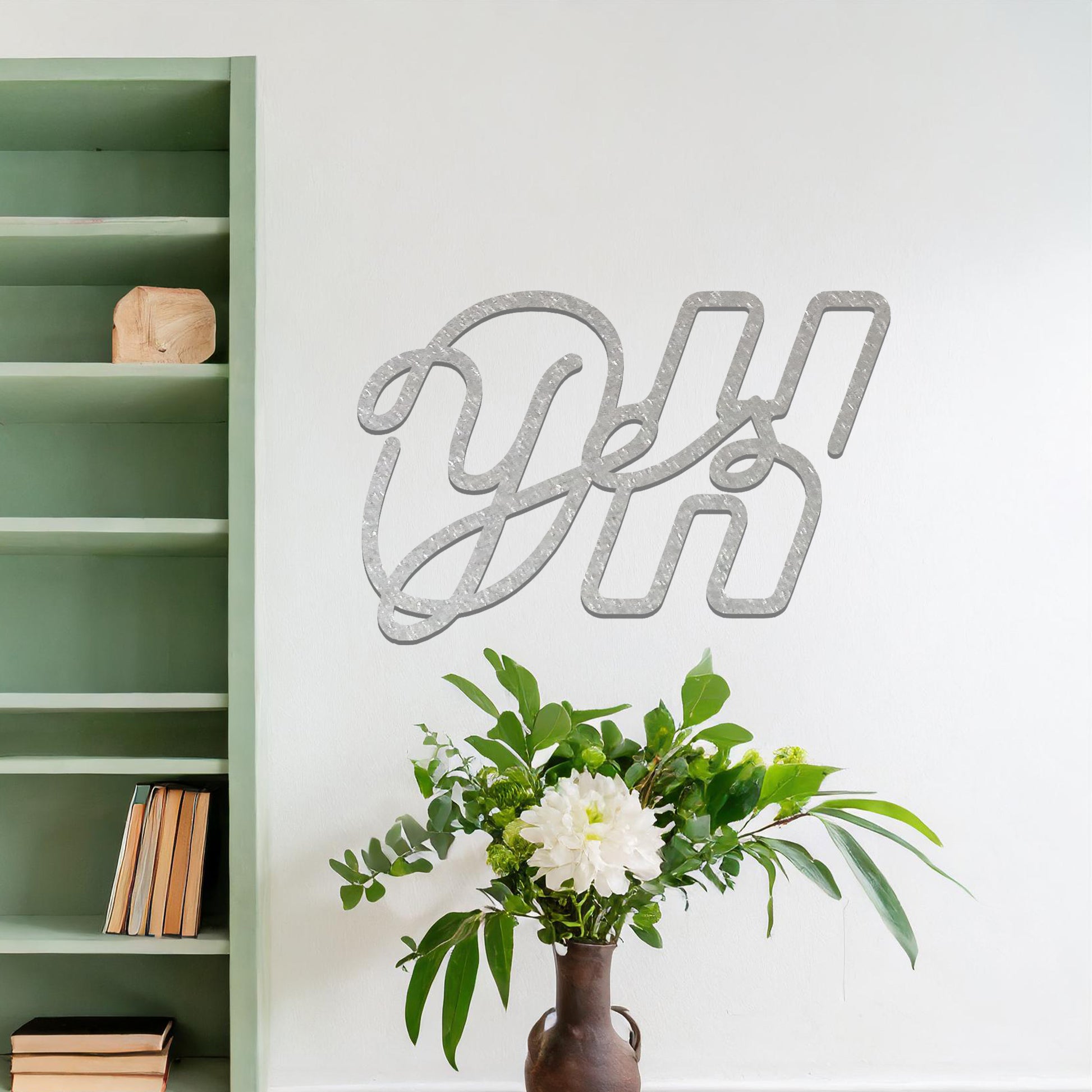 A contemporary metal wall art design featuring the bold, interwoven letters "Oh Yes" in a flowing, cursive style. The artwork is displayed on a neutral wall in a modern room setting.