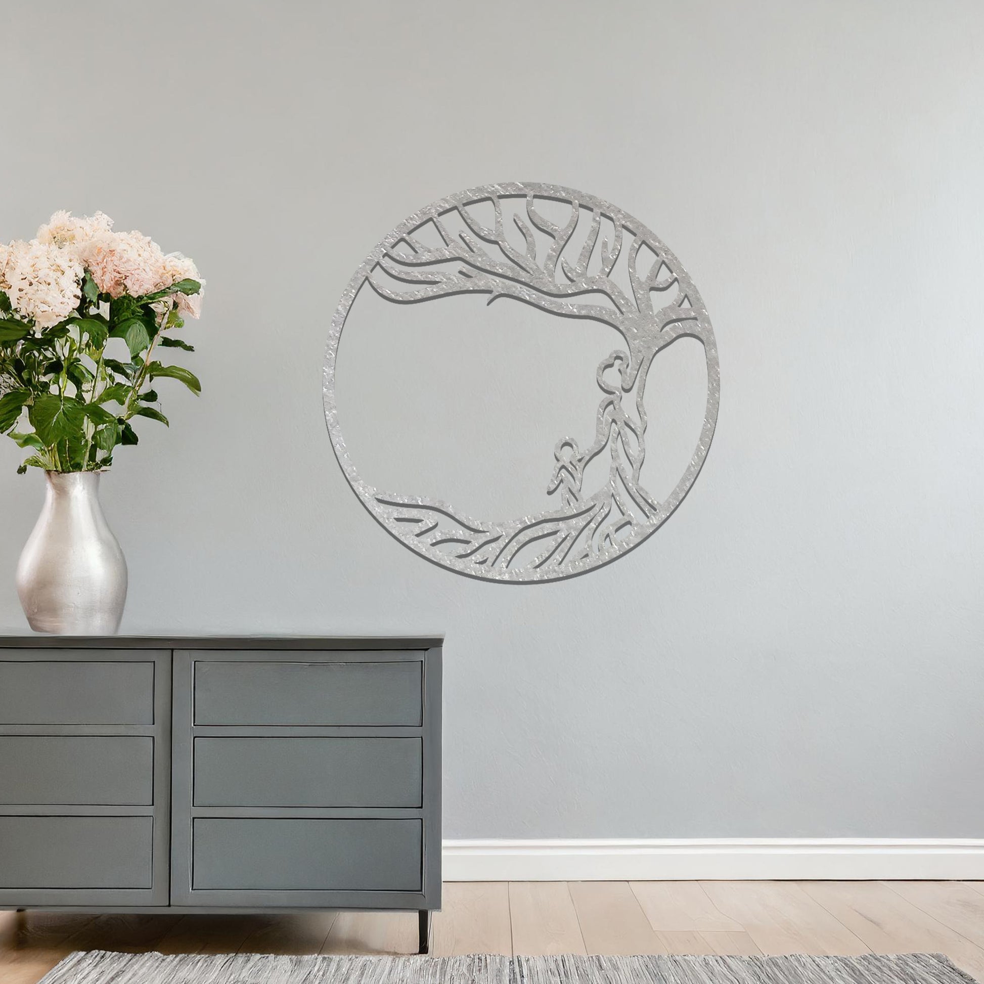 Metal wall art featuring a circular design with a symbolic tree and figures motif, depicting a parent and child under a tree with flowing branches, mounted on a neutral wall within modern room decor. The intricate design evokes warmth and connection, enhancing the aesthetic of the space.