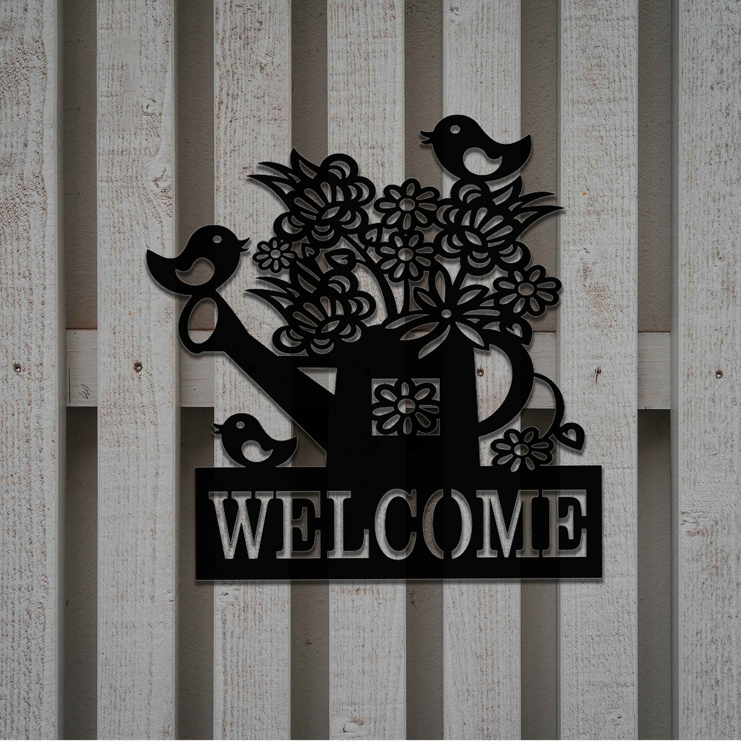Metal wall decoration featuring a watering can filled with flowers, birds perched on its handle, and the word "WELCOME" displayed below, crafted with intricate cutouts and a rustic finish, adding a warm and inviting touch to indoor or outdoor spaces.