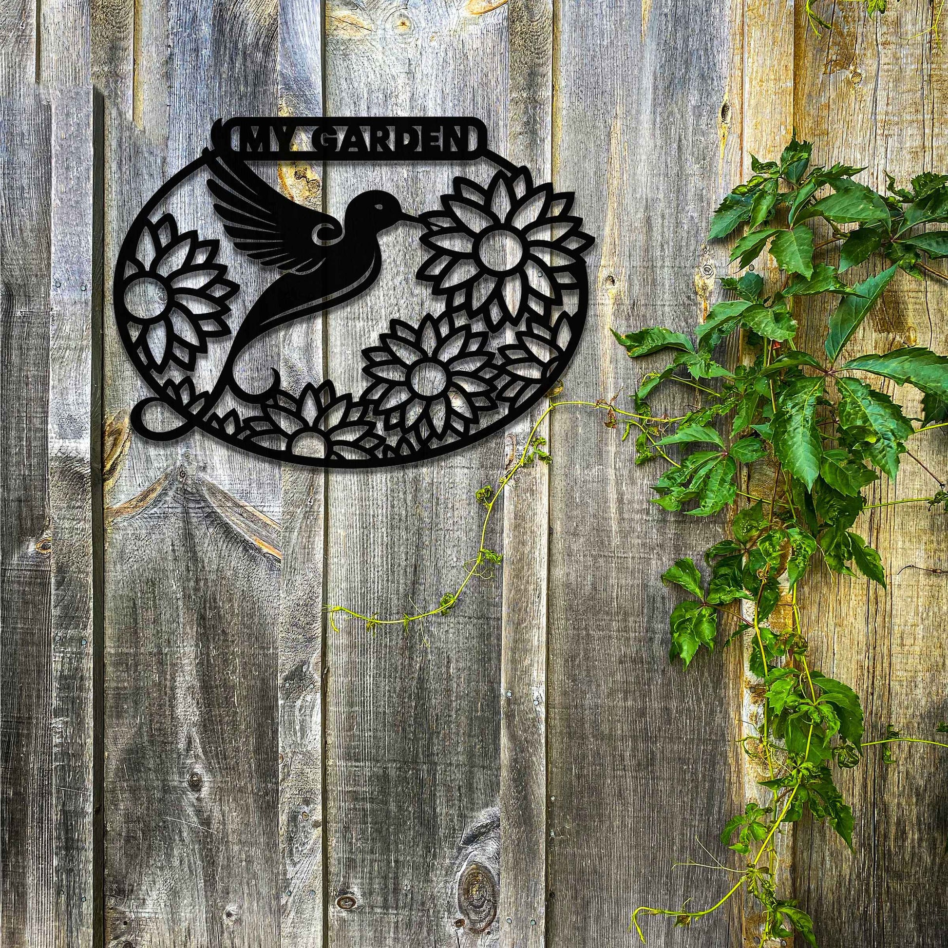 Metal garden decoration featuring a hummingbird surrounded by blooming sunflowers within an oval frame, inscribed with "My Garden," adding a personalised and nature-inspired charm to outdoor walls or fences.