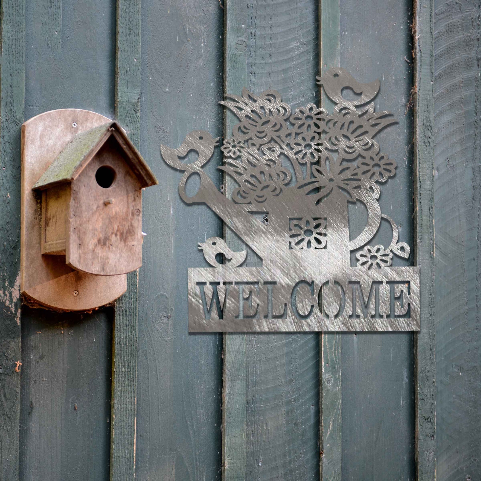 Metal wall decoration featuring a watering can filled with flowers, birds perched on its handle, and the word "WELCOME" displayed below, crafted with intricate cutouts and a rustic finish, adding a warm and inviting touch to indoor or outdoor spaces.