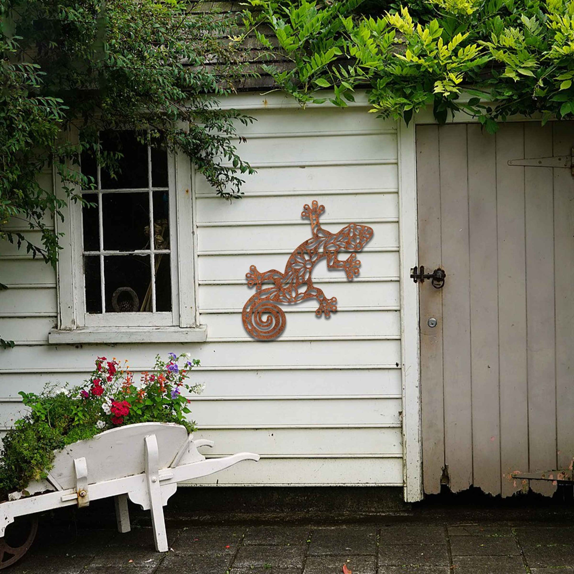 Metal garden decoration featuring a gecko design with floral and spiral cutouts, offering a playful and artistic accent to outdoor walls, seamlessly blending into rustic and natural settings.