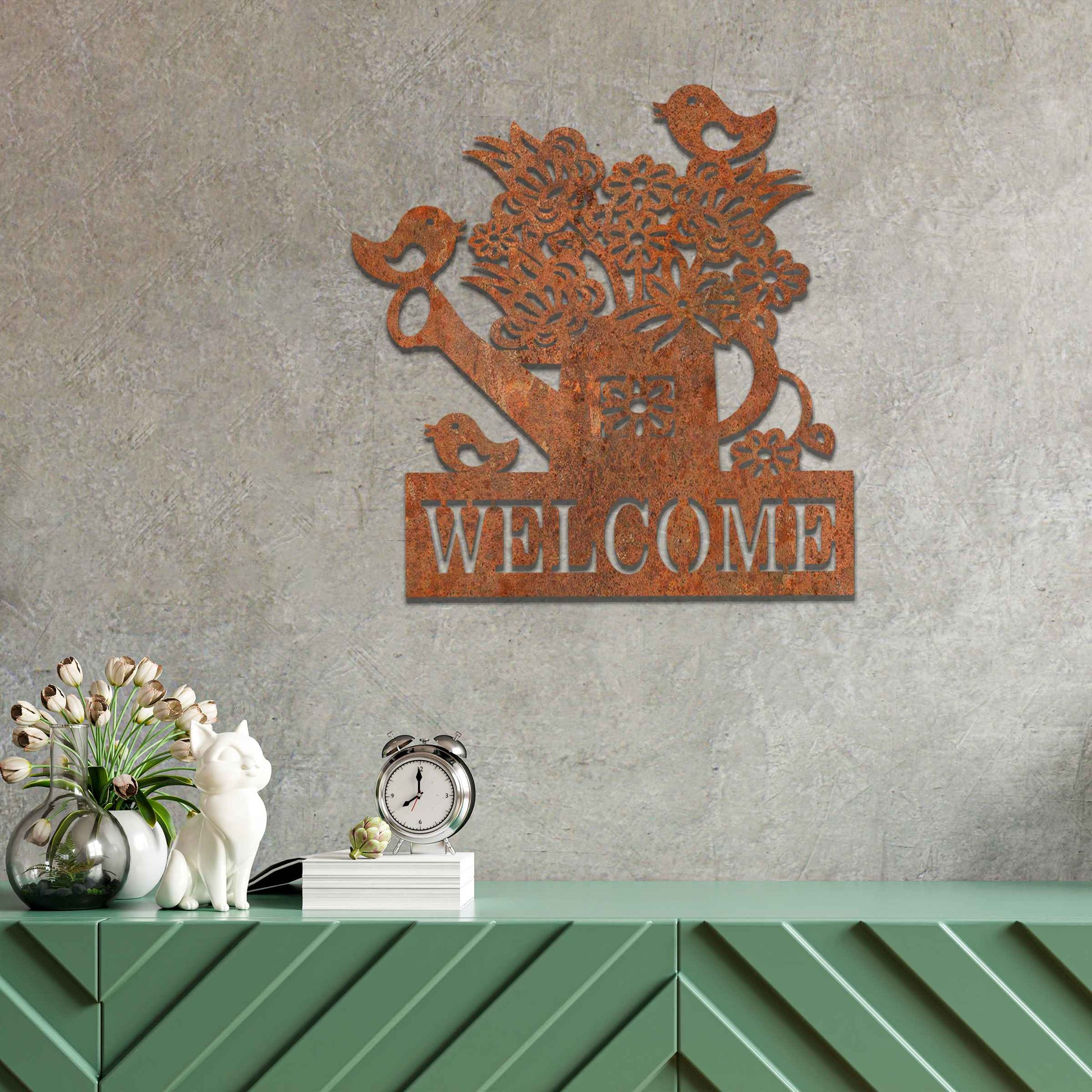 Metal wall decoration featuring a watering can filled with flowers, birds perched on its handle, and the word "WELCOME" displayed below, crafted with intricate cutouts and a rustic finish, adding a warm and inviting touch to indoor or outdoor spaces.