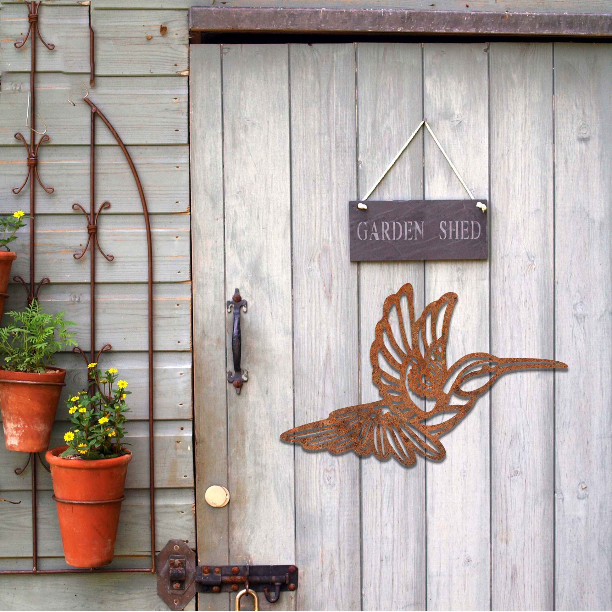 Metal garden decoration featuring a hummingbird in flight with intricate detailing, showcasing a rustic finish that adds a natural and artistic charm to outdoor walls or garden settings.