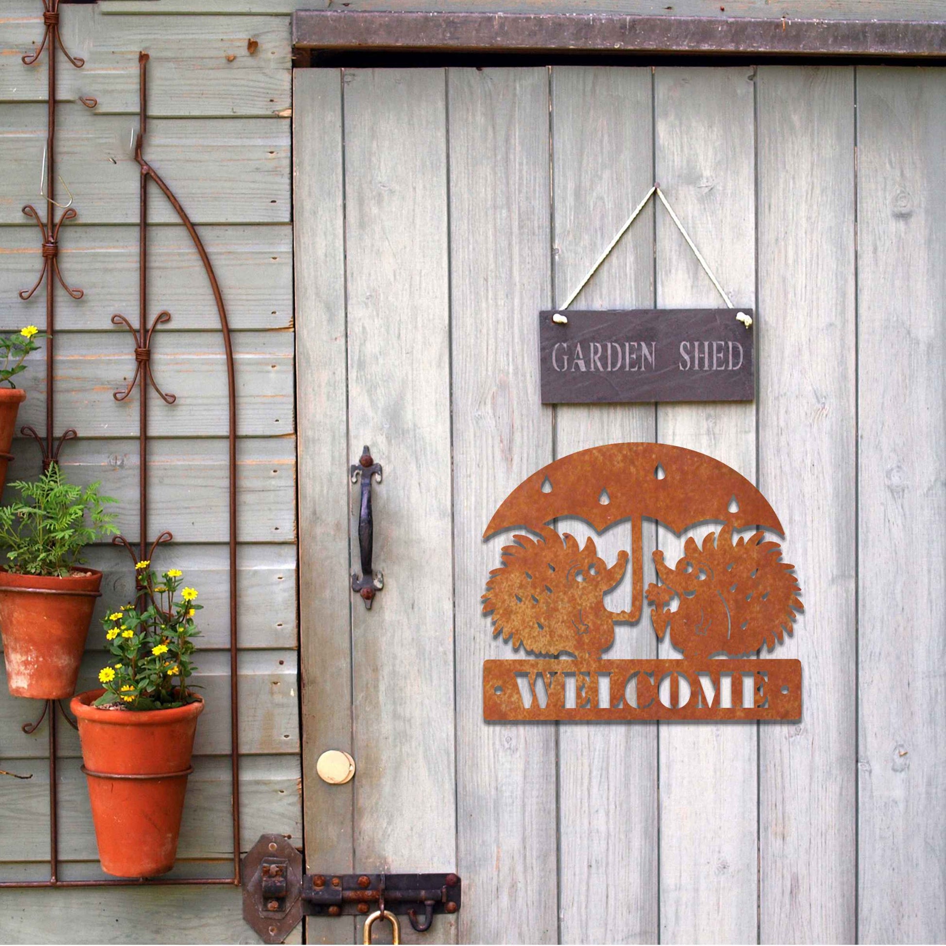 Metal garden decoration featuring two hedgehogs under an umbrella with raindrop details, accompanied by the word "Welcome," crafted with a rustic finish, adding a charming and inviting touch to outdoor spaces.