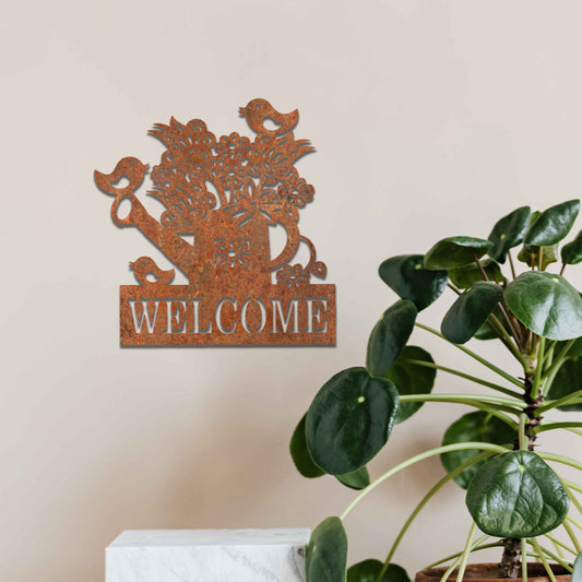 Metal wall decoration featuring a watering can filled with flowers, birds perched on its handle, and the word "WELCOME" displayed below, crafted with intricate cutouts and a rustic finish, adding a warm and inviting touch to indoor or outdoor spaces.