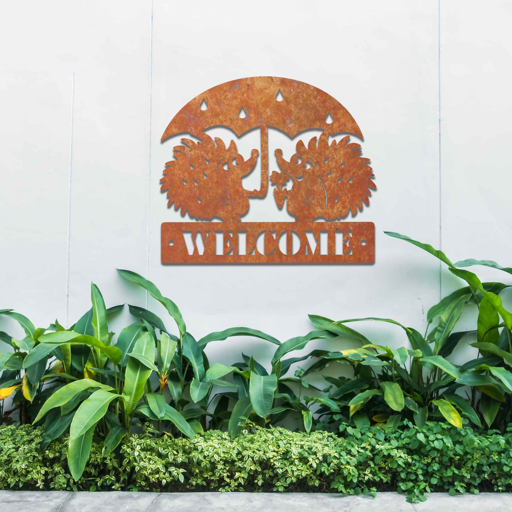 Metal garden decoration featuring two hedgehogs under an umbrella with raindrop details, accompanied by the word "Welcome," crafted with a rustic finish, adding a charming and inviting touch to outdoor spaces.