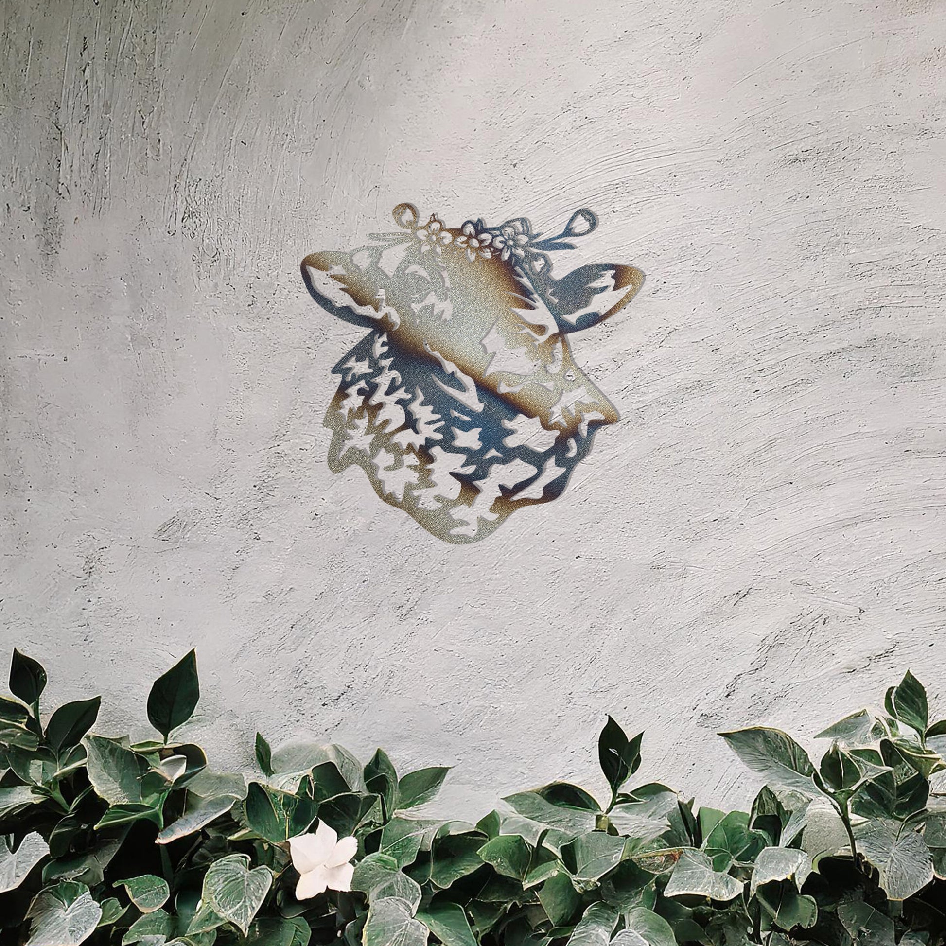 A metal wall art piece depicting a sheep adorned with a floral crown, exuding rustic charm and elegance. This intricate design brings a touch of countryside serenity to any modern or natural setting, complemented by its leafy surroundings.