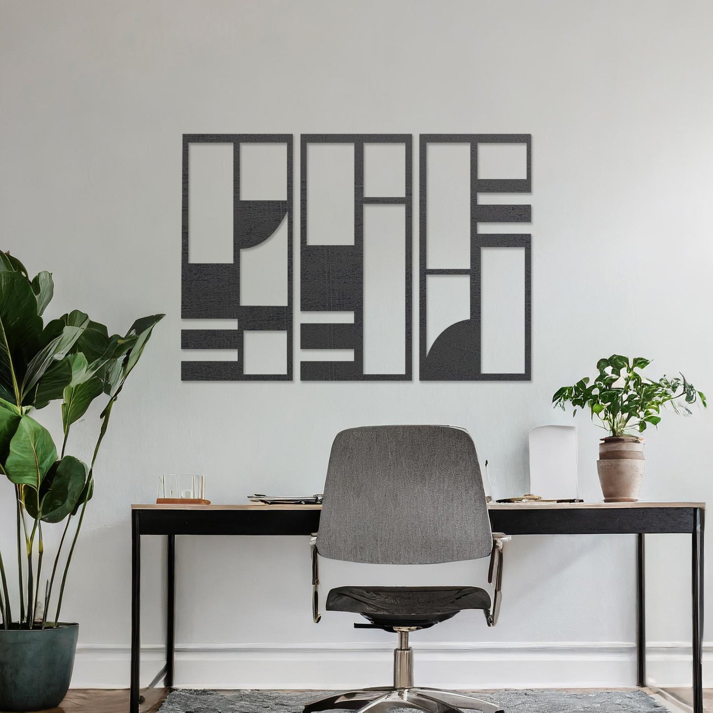 This image displays a modern, abstract triptych wall art piece featuring geometric patterns in dark tones. The artwork is placed above a simple desk setup with a plant and a chair, providing a stylish and minimalistic atmosphere suitable for a contemporary workspace. 