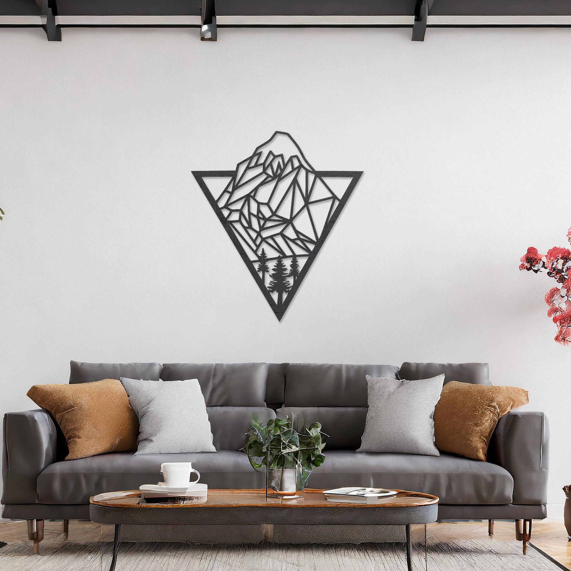 Triangular wall art featuring a geometric mountain landscape design with trees at the base, evoking nature and adventure. The piece is displayed on a neutral wall in a modern living room with a stylish sofa and contemporary decor.