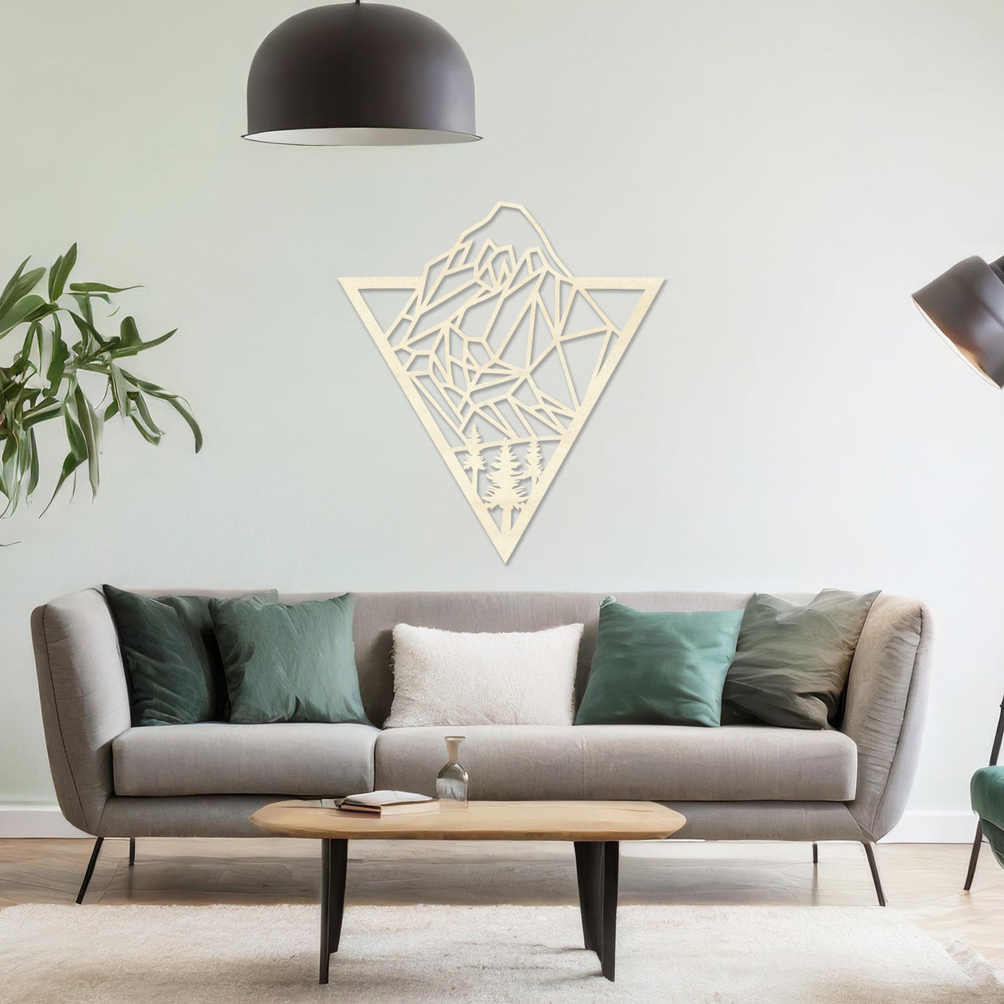 Triangular wall art featuring a geometric mountain landscape design with trees at the base, evoking nature and adventure. The piece is displayed on a neutral wall in a modern living room with a stylish sofa and contemporary decor.