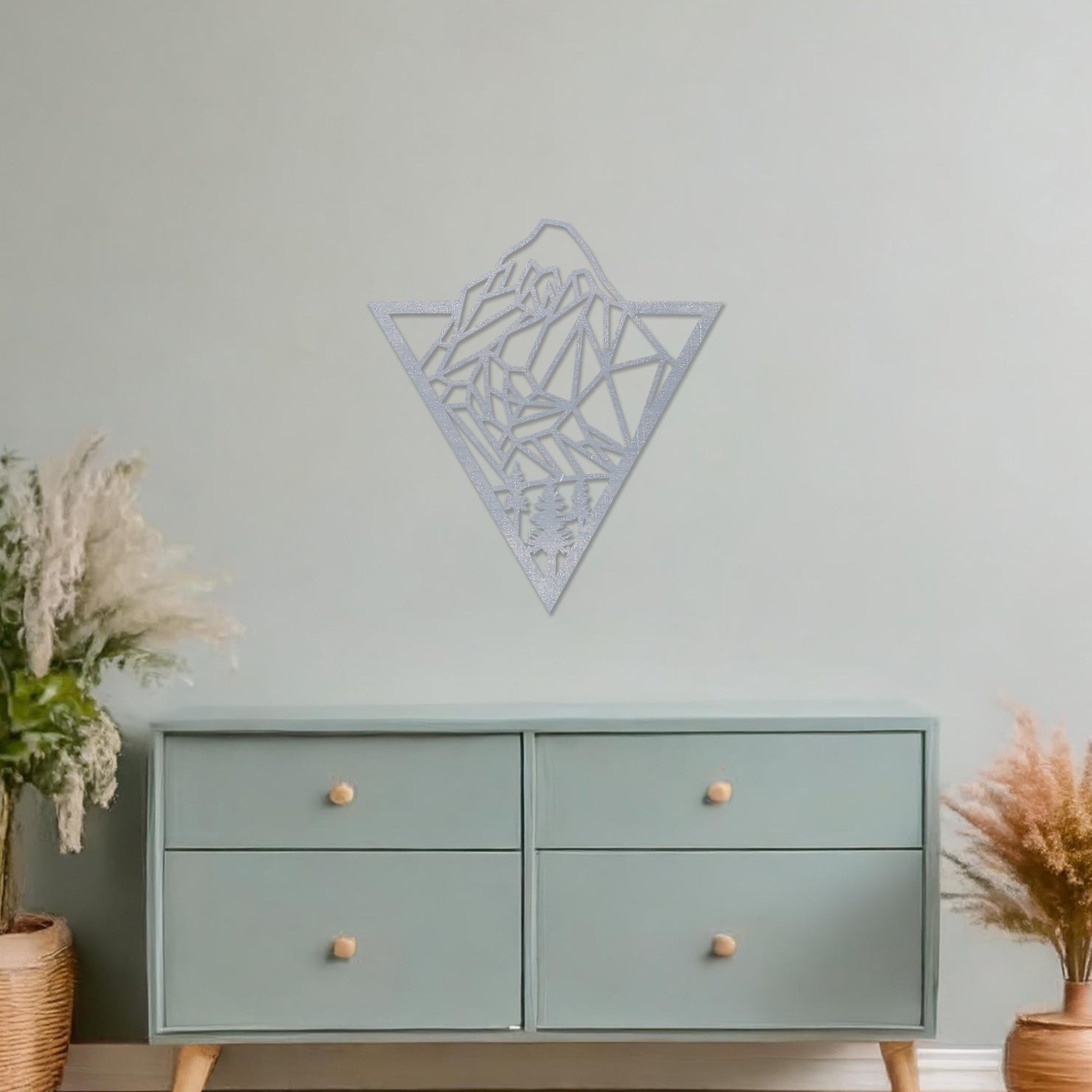 Triangular wall art featuring a geometric mountain landscape design with trees at the base, evoking nature and adventure. The piece is displayed on a neutral wall in a modern living room with a stylish sofa and contemporary decor.